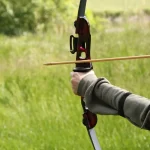 close-up of a hand holding a bow and aiming at a target, in the background a green landscape, a boy performs archery