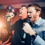 two young caucasian men in t-shirts singing in microphone in karaoke bar, having fun, celebrating. holiday, leisure, party concept